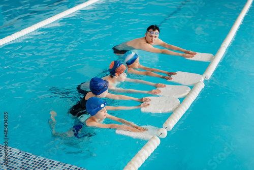 A group of boys and girls train and learn to swim in a modern swimming pool with an instructor. Development of children s sports. Healthy parenting and promotion of children s sports.