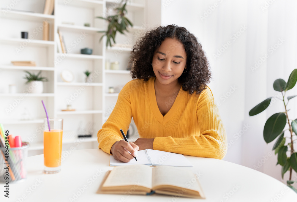 Young black woman writing in notebook, getting ready for exam or test, sitting at desk indoors Young black woman writing in notebook, getting ready for exam or test, sitting at desk indoors