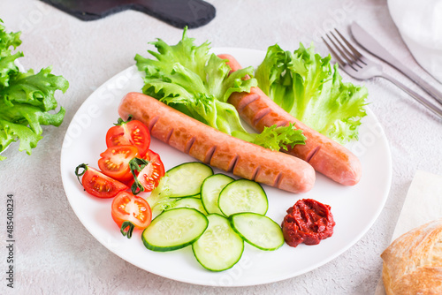 Ready to eat fried sausages, chopped tomatoes and cucumber vegetables on a plate and a bun on the table. Hearty homemade snack