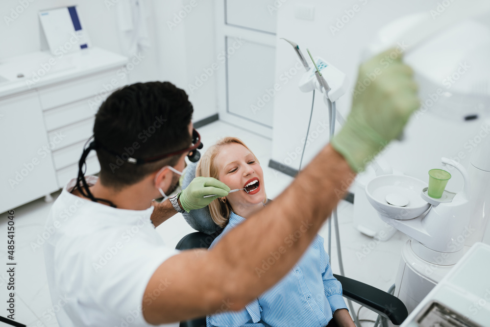 Fototapeta premium Beautiful young woman having dental treatment at dentist's office. Dentist is wearing protective face mask due to coronavirus pandemic..