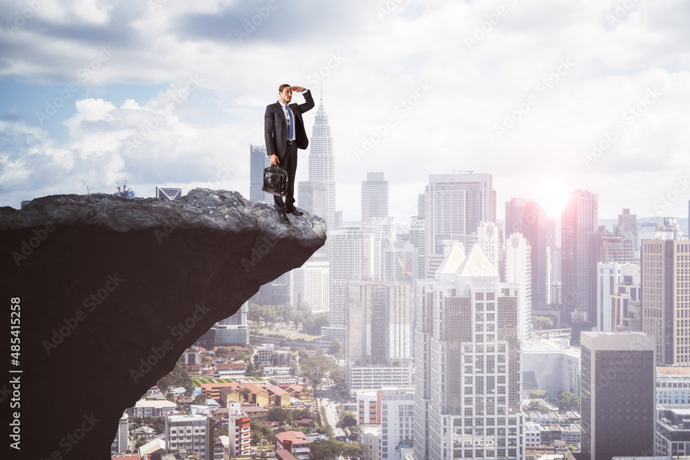 Handsome young european businessman on cliff edge looking into the distance on bright city background with daylight and mock up place. Future, success, leadership and career growth concept. Handsome young european businessman on cliff edge looking into the distance on bright city background with daylight and mock up place. Future, success, leadership and career growth concept.