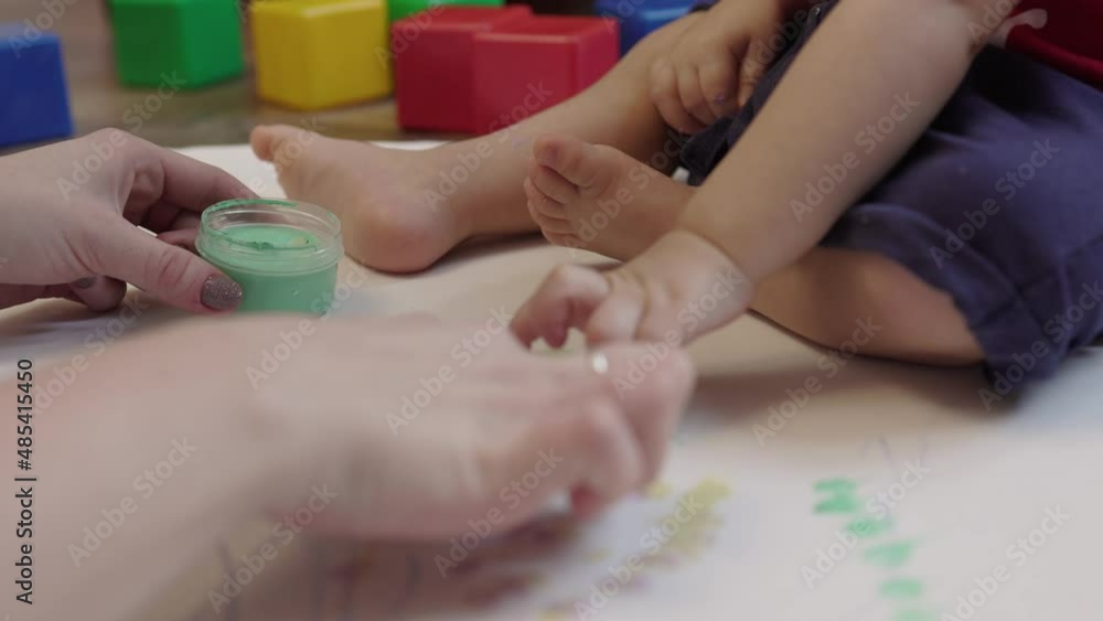 mother teaches toddler baby painting with finger paints on white paper. child sitting on floor taking paint bottle in hands mom showing how put finger in paint and color paper. kids home education mother teaches toddler baby painting with finger paints on white paper. child sitting on floor taking paint bottle in hands mom showing how put finger in paint and color paper. kids home education