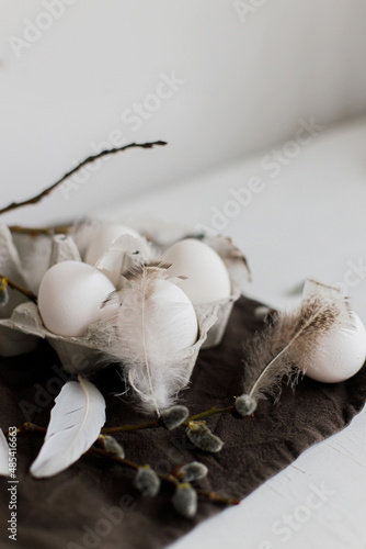 Stylish Easter still life. Natural eggs in tray, feathers, willow branches on rustic linen napkin on white wooden table. Simple rural Easter aesthetics in pastel white and brown colors