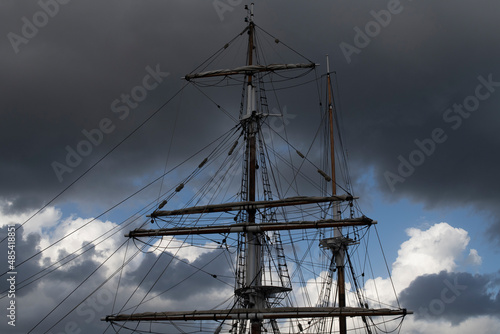 Top of ship mast against a clearing sky. Dramatic dark clouds frame the mast's tip and a window of blue sky in the centre. Fluffy white and grey clouds beneath blue sky.