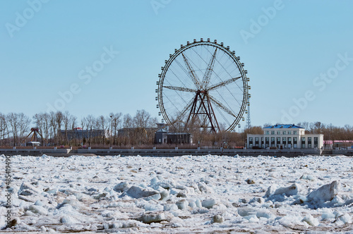 Ferris wheel on the bank of the icy Amur River on a spring day. The beginning of the ice drift. Embankment of Heihe Island, China. View from the city of Blagoveshchensk, Russia. Sunny day.