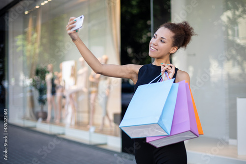 shopping, sale, technology and people concept - Portrait of a young African-American girl with colorful bags taking selfies on her phone outdoors in front of the mall