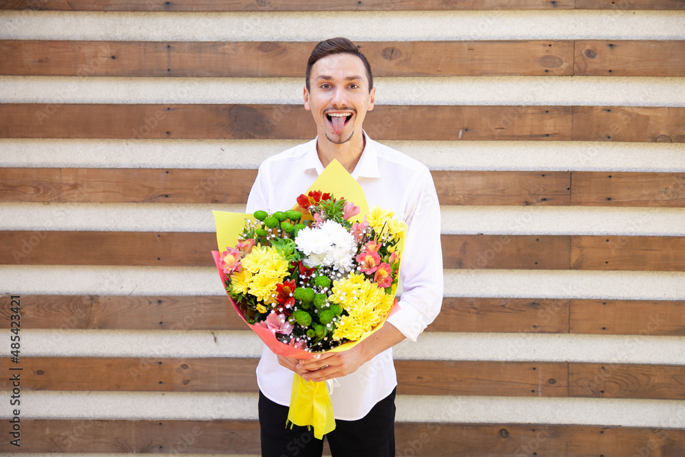 Fototapeta premium Summer, flowers, emotions and people concept - Satisfied Caucasian guy in a white shirt and black jeans showing tongue and holding a big bouquet on brown ribbed background with copy space