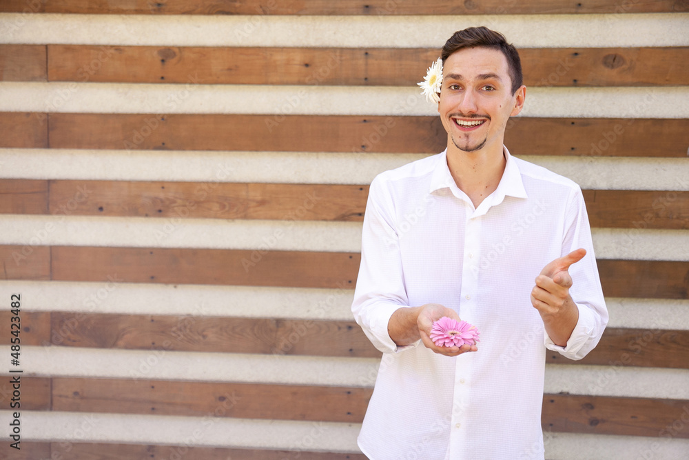 Obraz premium emotions, gesture, blossoms and people concept - Smiling guy with a camomile behind his ear, holding a pink flower in his hands and pointing finger at the camera on a ribbed background with copy space
