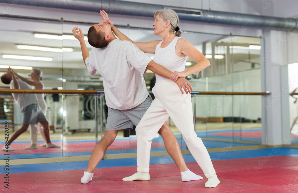 Obraz premium Focused elderly woman practicing basic self-defense techniques while training in gym with male partner, performing palm heel strike in chin