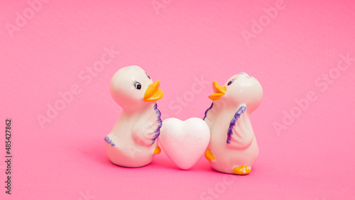 two ceramic ducks and white heart on pink background. Symbols of love and fidelity. Cute greeting card for wedding invitation, for Valentine's day. Illustration of the feeling of love between two.