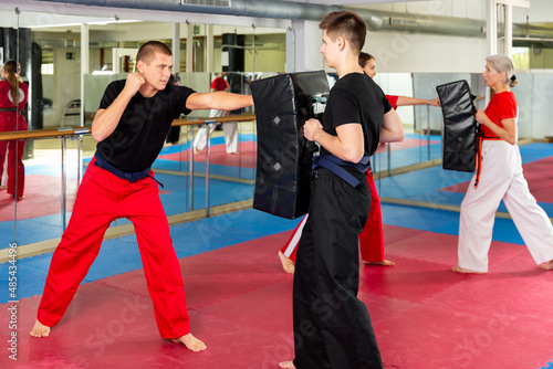 Caucasian man exercising jabs with teenager boy who holding punching pad during group karate training.
