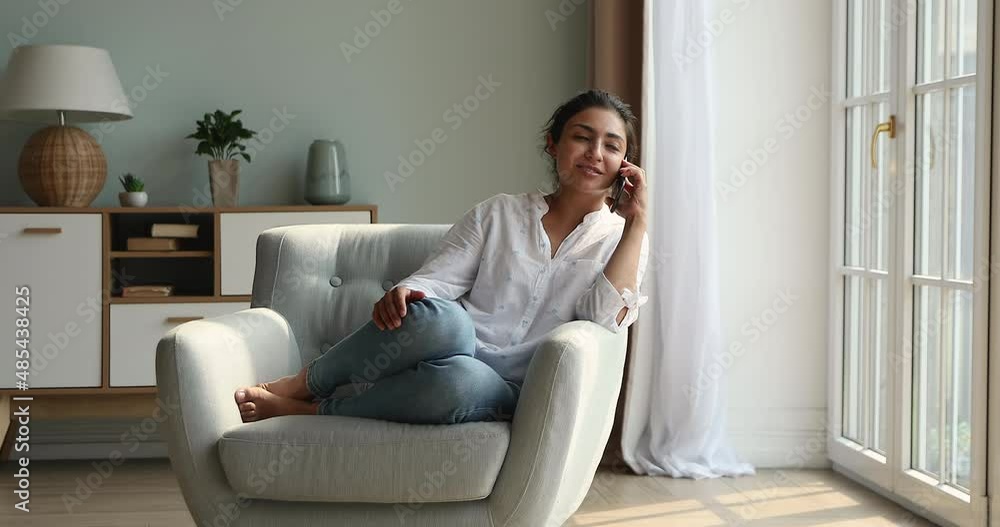 Cute Indian woman relaxing sit on armchair in cozy living room holds cellphone talking to friend, lead personal conversation on smartphone, having distancing communication. Tech, phone call concept Cute Indian woman relaxing sit on armchair in cozy living room holds cellphone talking to friend, lead personal conversation on smartphone, having distancing communication. Tech, phone call concept