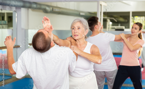 Determined aged woman practicing effective self defence techniques with male sparring partner in training room, using palm to launch blow in chin