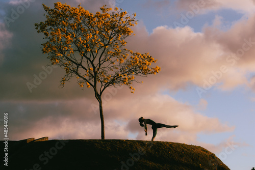 Beautiful health woman silhouette in pink blue sky sunrise doing yoga at the hill with a yellow tree