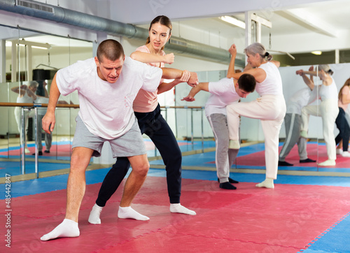 Caucasian woman performing elbow strike while sparring with man in gym during self-defence training. Senior woman training in background.