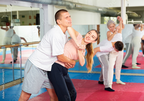 European woman learning elbow strike move during self-defense training. Mature woman training with sparring-partner in background.