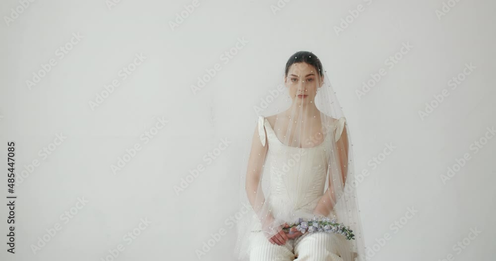 Gorgeous young female posing with flowers in her hands and looking seductively at camera sitting on clean background. Wedding photo session of charming sensual bride under veil. Pretty stylish woman Gorgeous young female posing with flowers in her hands and looking seductively at camera sitting on clean background. Wedding photo session of charming sensual bride under veil. Pretty stylish woman
