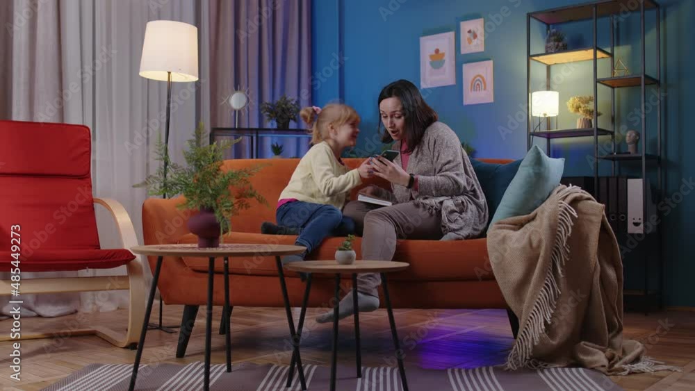 Little child kid daughter girl ran up to mother who is reading a book and showing smartphone display sincerely rejoicing win, receiving message with good news, success luck together at home on sofa Little child kid daughter girl ran up to mother who is reading a book and showing smartphone display sincerely rejoicing win, receiving message with good news, success luck together at home on sofa