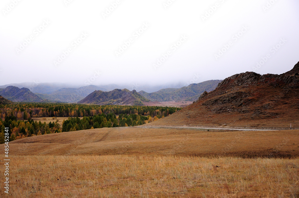 An asphalt road skirting a high hill goes through a dry autumn steppe under thunderclouds.