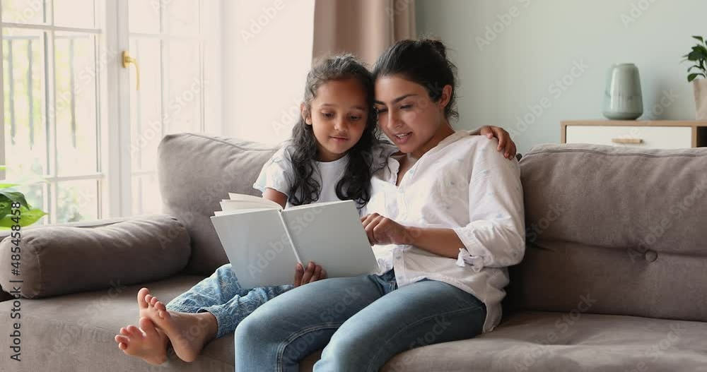 Cheerful Indian woman read fairytale book to her cute little daughter sit together on couch at home. 6s girl enjoy fascinating story, listen mommy storytelling. Kid development and upbringing concept Cheerful Indian woman read fairytale book to her cute little daughter sit together on couch at home. 6s girl enjoy fascinating story, listen mommy storytelling. Kid development and upbringing concept