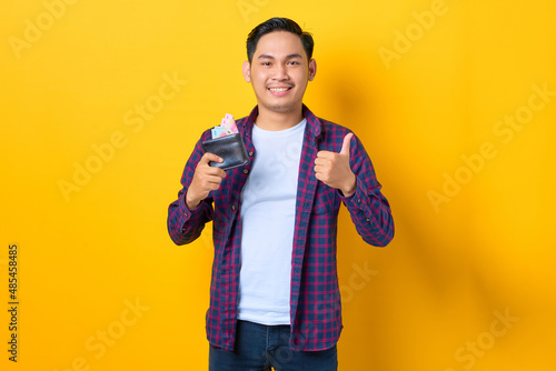 Smiling young Asian man in plaid shirt holding wallet full of money banknotes and showing thumb up isolated on yellow background