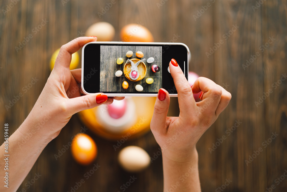 Woman holding mobile phone and making photo of eggs painted for Easter isolated on wooden background. Easter eggs flat lay on rustic table. Easter concepts