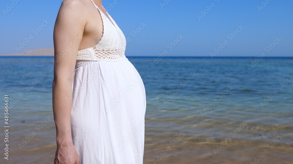 Beauty of pregnancy period, woman with big baby bump in white dress walking on the sunny ocean coast Beauty of pregnancy period, woman with big baby bump in white dress walking on the sunny ocean coast