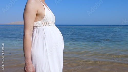 Beauty of pregnancy period, woman with big baby bump in white dress walking on the sunny ocean coast 