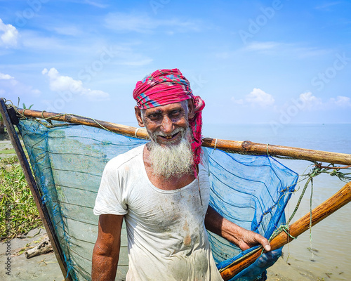 Village fisherman portrait with a lave net in hand, rural old man is standing beside a river, Bangladeshi fisherman with white dress and towel in head. photo