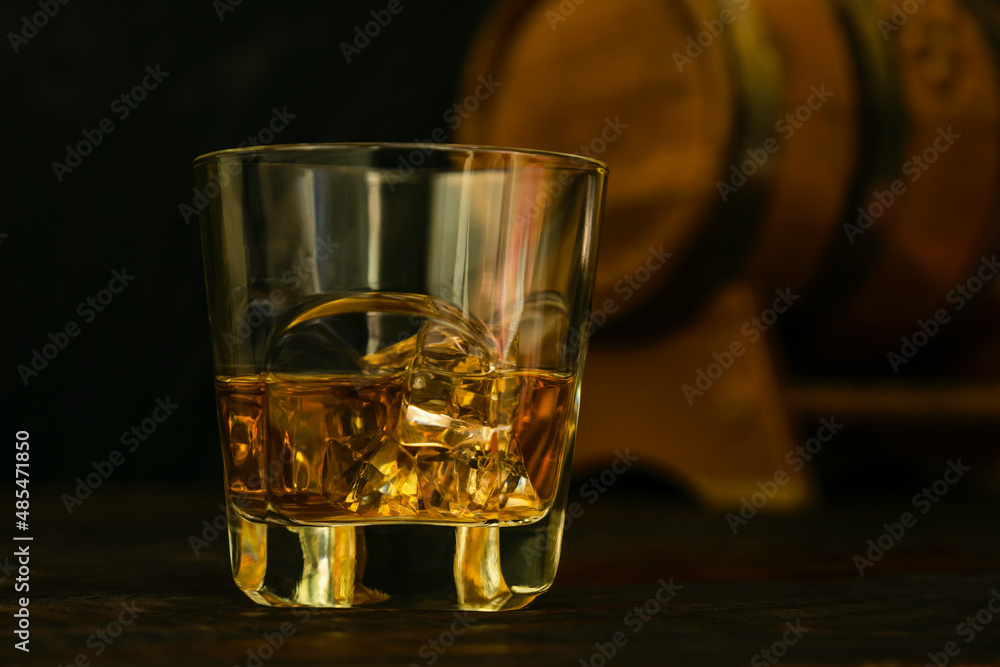 A glass of whiskey with ice cubes against the background of an oak barrel. Close-up. Dark background. A glass of whiskey with ice cubes against the background of an oak barrel. Close-up. Dark background.