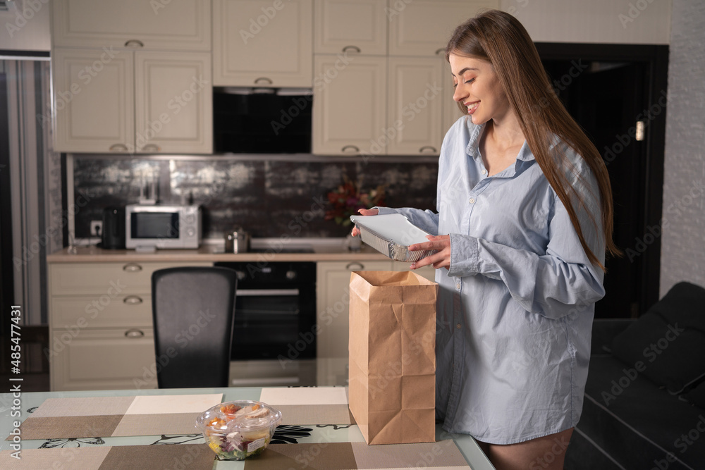 A beautiful Caucasian girl at home in the evening at home takes disposable lunch boxes with food from a paper bag and puts them on the table. Takeaway food concept. A beautiful Caucasian girl at home in the evening at home takes disposable lunch boxes with food from a paper bag and puts them on the table. Takeaway food concept.