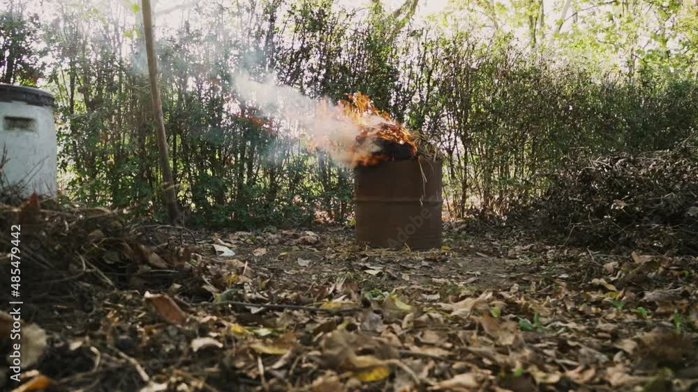 A man in dirty clothes adjusts and pokes the smoking green waste dumpster fire in the garden with a fork, slow motion locked shot A man in dirty clothes adjusts and pokes the smoking green waste dumpster fire in the garden with a fork, slow motion locked shot