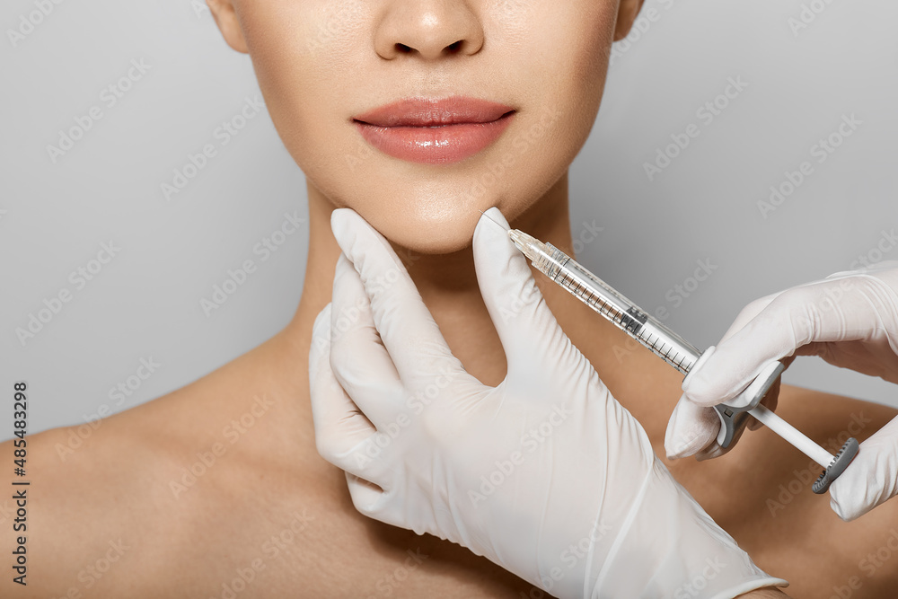 Syringe near woman's chin, beauty injections with fillers for chin shape correction. Cropped female face while procedure chin contouring - obrazy, fototapety, plakaty 