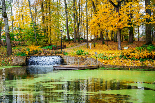 Small cascading waterfall with a jetty on the shore of a quiet lake on the background an autumn foliage forest in Latvia. oncept of a relaxing weekend holiday in nature.