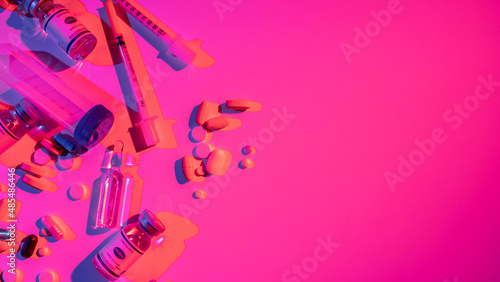 Medication banner. Antiviral treatment. Coronavirus immunization. Neon light vaccine ampoule dose syringe capsules test tube scattered on bright pink color empty space background.
