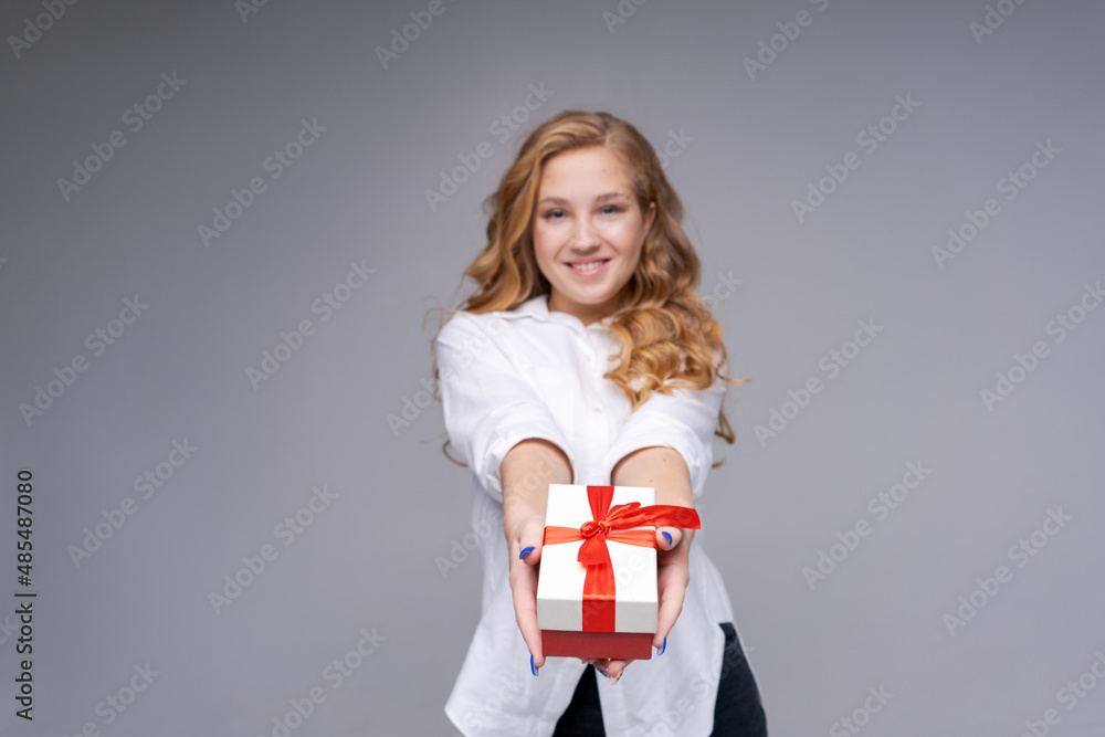 Portrait an attractive happy funny girl holding gift box on light gray background in a white shirt with long wavy hair, copy space. Birthday gift. Women's day, march 8 Portrait an attractive happy funny girl holding gift box on light gray background in a white shirt with long wavy hair, copy space. Birthday gift. Women's day, march 8