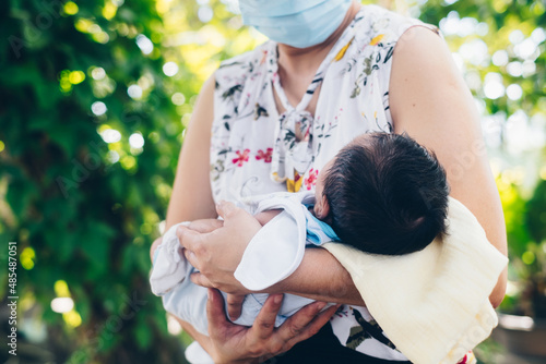 Unrecognizable woman wearing a disposable face mask holding a month old baby boy in her arms. Closeup. Copy space.