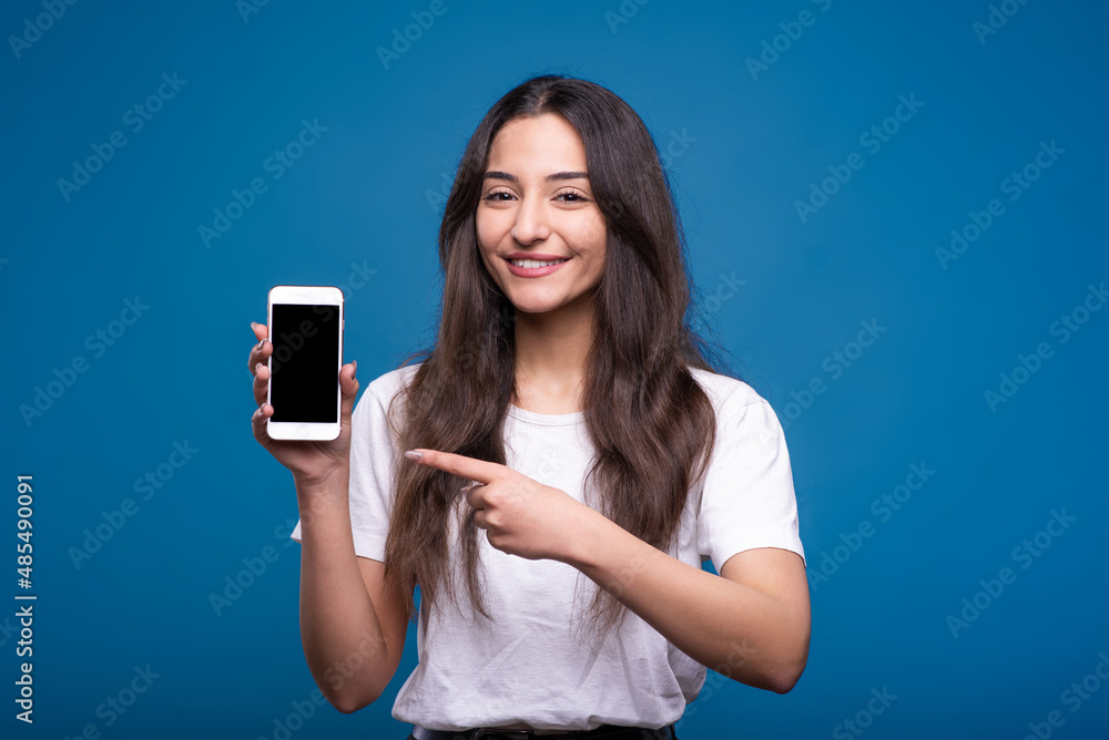 Attractive caucasian or arab brunette girl in white t-shirt pointing finger at smartphone screen with copy space for advertising isolated on blue studio background. Attractive caucasian or arab brunette girl in white t-shirt pointing finger at smartphone screen with copy space for advertising isolated on blue studio background.