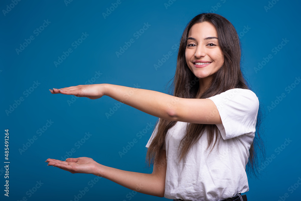 Attractive caucasian or arab brunette girl in a white t-shirt showing a big object with her hands on a copy space for advertising isolated on a blue studio background. Attractive caucasian or arab brunette girl in a white t-shirt showing a big object with her hands on a copy space for advertising isolated on a blue studio background.