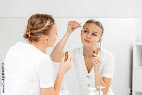 A young woman looks in the mirror and applies cosmetic oil on her face. Back view from the shoulder. Skin care concept
