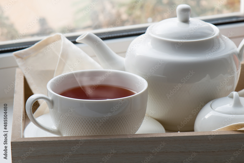 A white cup with hot tea on a saucer, a teapot, a sugar bowl and a cloth napkin on a wooden tray against the background of the window. Close-up. A white cup with hot tea on a saucer, a teapot, a sugar bowl and a cloth napkin on a wooden tray against the background of the window. Close-up.