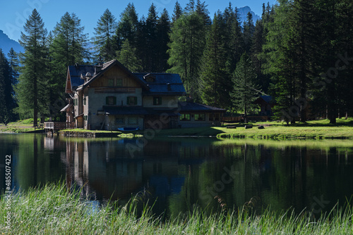 early in the morning  a house by a mountain lake in the Alps surrounded by Christmas trees in the background high Alps  blue sky. Reflections of trees and mountains in the water