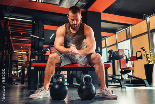 A young bearded athletic man trains in the gym with dumbbells. The athlete rubs magnesia in his hands. The concept of fitness
