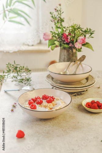 White bowl with vegan porridge with raspberries at white concrete kitchen table with crockery and flower bunch at window background with natural light. Healthy breakfast at home. Front view.