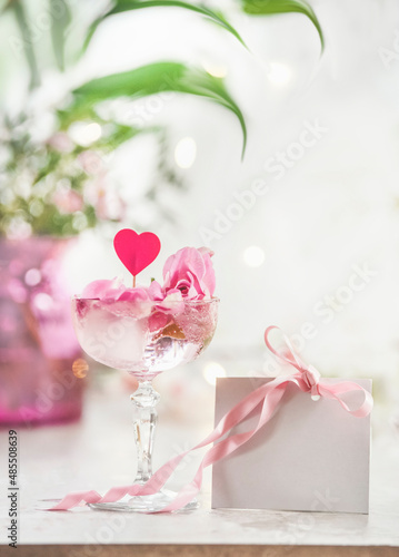 Champagne glass with pink rose petals, heart and blank white gift card with pink ribbon on white table at floral bokeh background. Celebrating valentines day. Front view