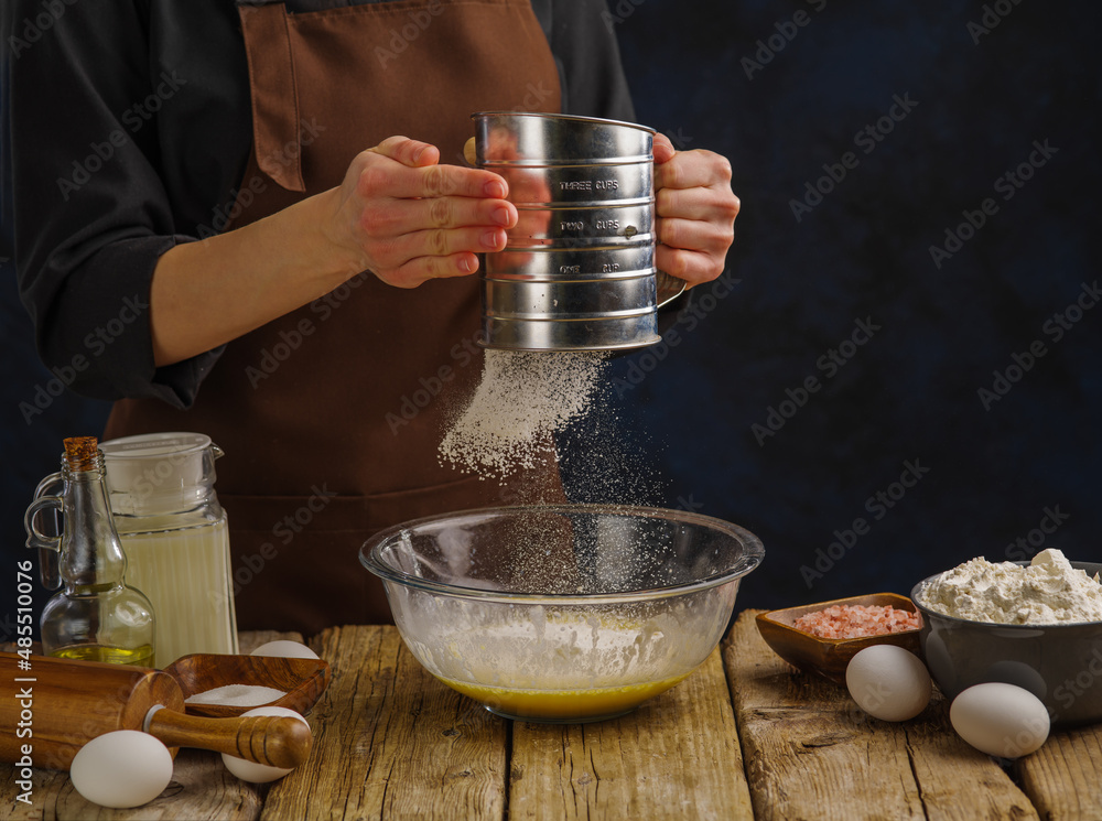 The chief is sifting flour. Prepares the dough on a wooden table and a dark background. Preparation of dishes from dough - pizza, bread, pasta, pies. Advertising, banner, culinary blog. The chief is sifting flour. Prepares the dough on a wooden table and a dark background. Preparation of dishes from dough - pizza, bread, pasta, pies. Advertising, banner, culinary blog.