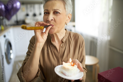 Pretty stylish grey haired mature Caucasian woman blowing gold festive horn  holding plate with cup cake in hand getting ready to make birthday surprise for her husband returning home from work