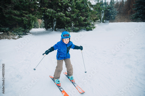 Little boy with skis, runs on the ski slope in the forest. Child with ski equipment smiles and has fun on the slope. Child in ski school learns to ski in Germany, France or Poland. 