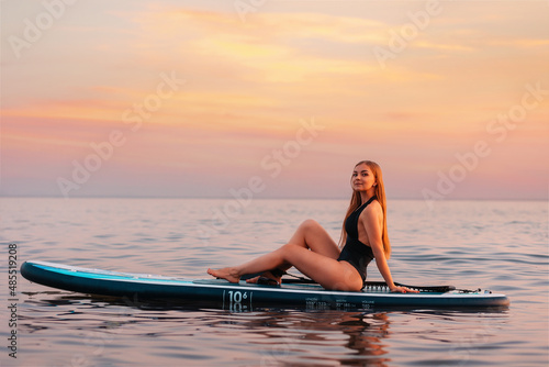 Surfing. Elegant woman sitting on a sup board at ocean surface. In the background, the ocean and the sunset. Summer activity
