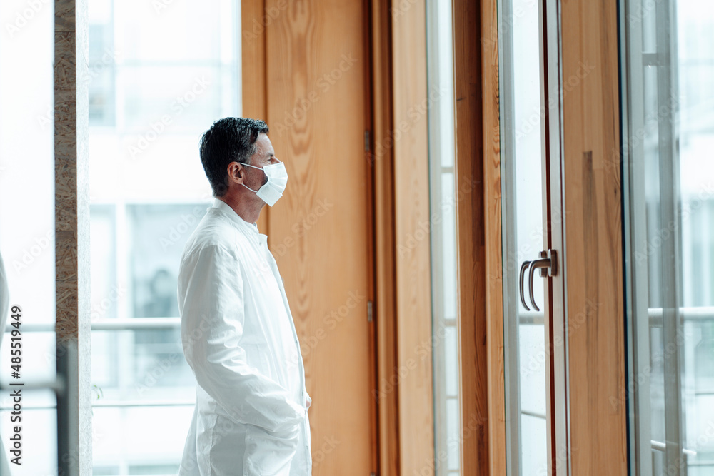 Scientist with hand in pocket looking out of window wearing protective face mask at clinic Scientist with hand in pocket looking out of window wearing protective face mask at clinic
